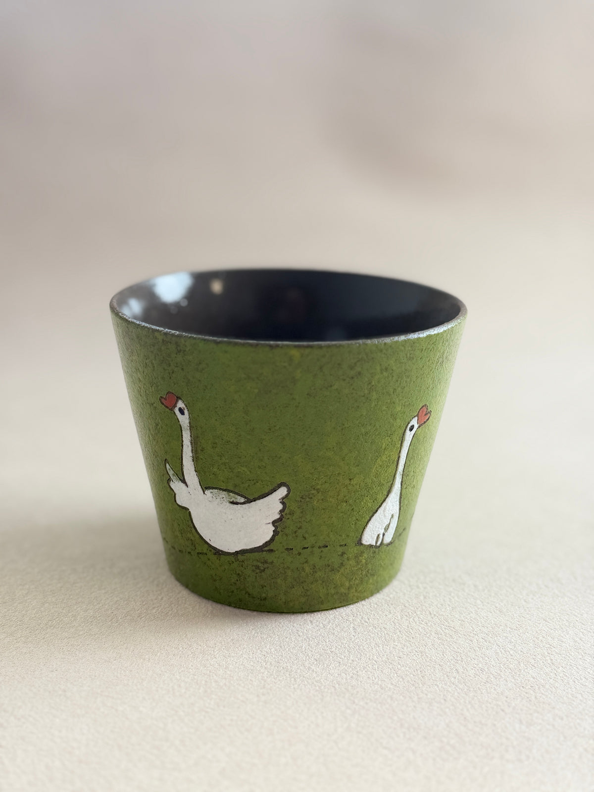 A small speckled olive green ceramic cup with a dark navy blue interior, decorated with two white swans facing each other. The cup is photographed on a neutral, light beige textured surface.