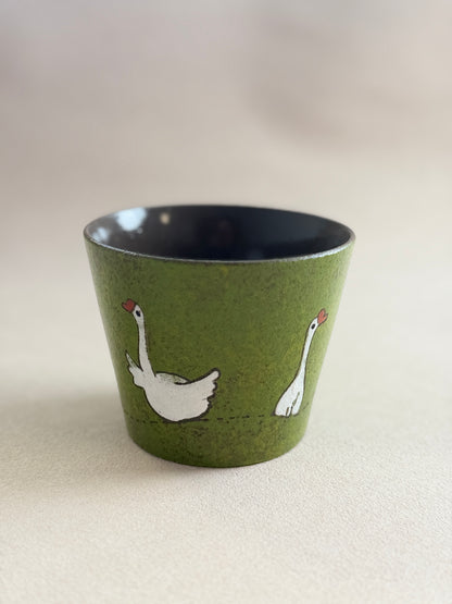 A small speckled olive green ceramic cup with a dark navy blue interior, decorated with two white swans facing each other. The cup is photographed on a neutral, light beige textured surface.