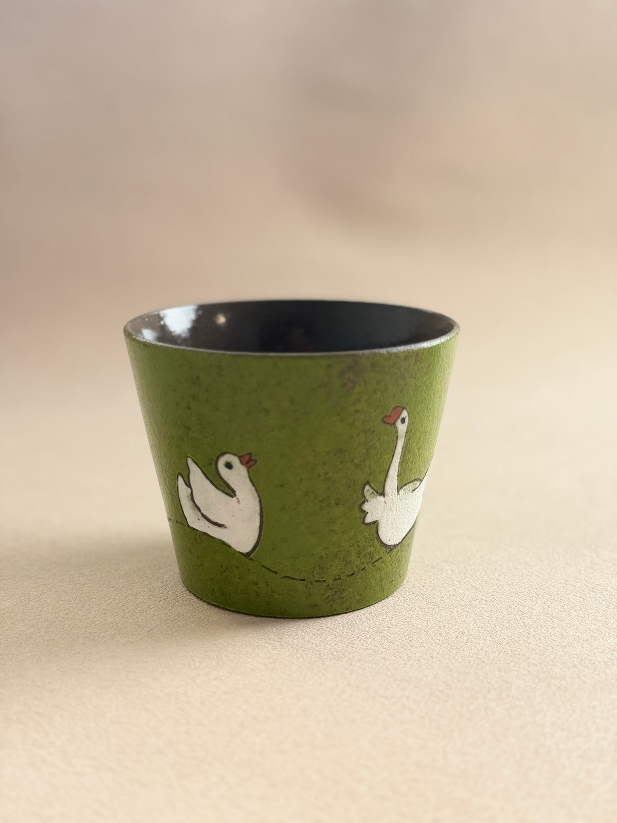 A small green ceramic cup with a black interior, decorated with two white swans facing each other, set against a plain beige background.