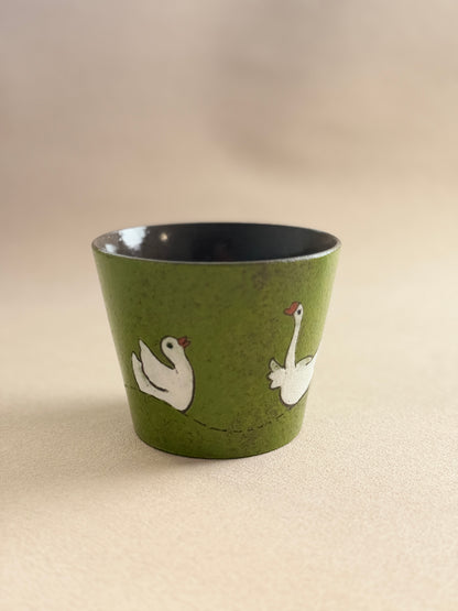 A small green ceramic cup with a black interior, decorated with two white swans facing each other, set against a plain beige background.