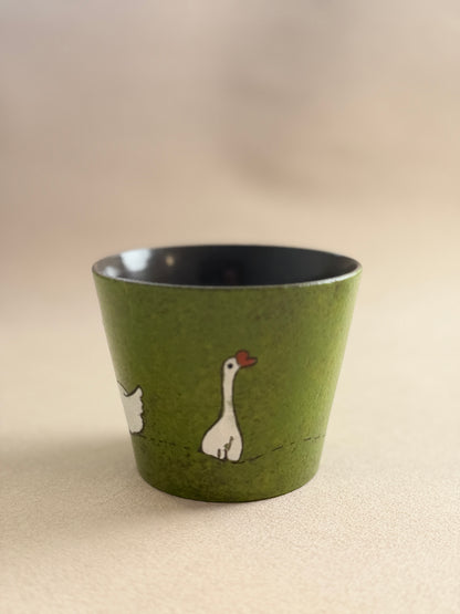 A small green ceramic cup with a hand-painted white goose illustration and a red beak, set against a warm beige background.