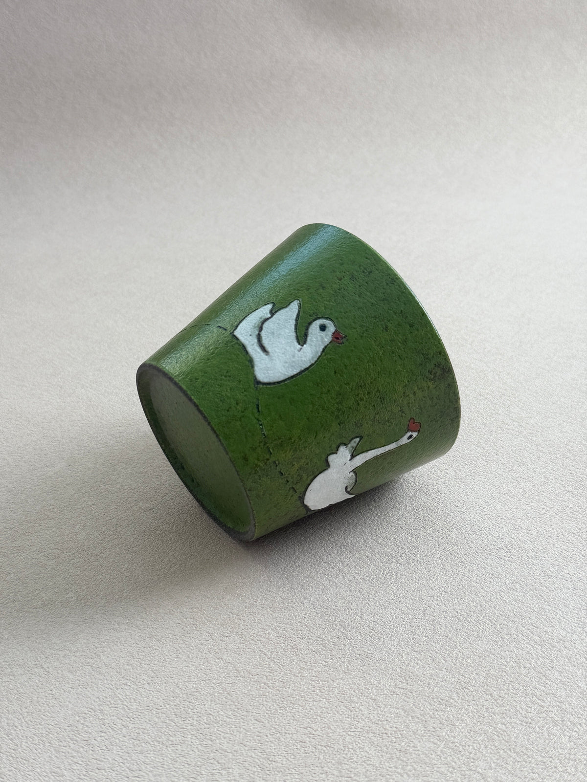 A green ceramic cup with hand-painted white swans on a textured beige background.
