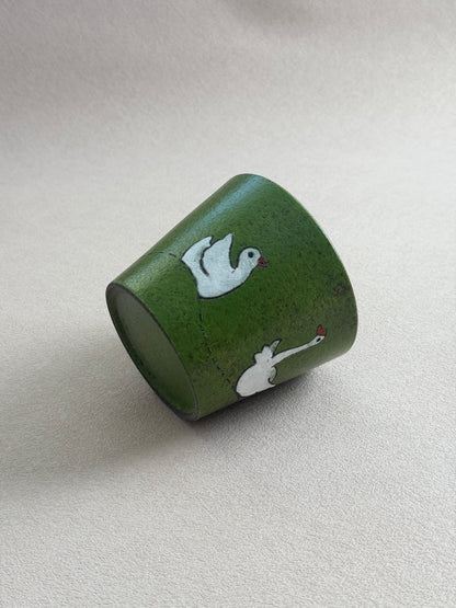 A green ceramic cup with hand-painted white swans on a textured beige background.