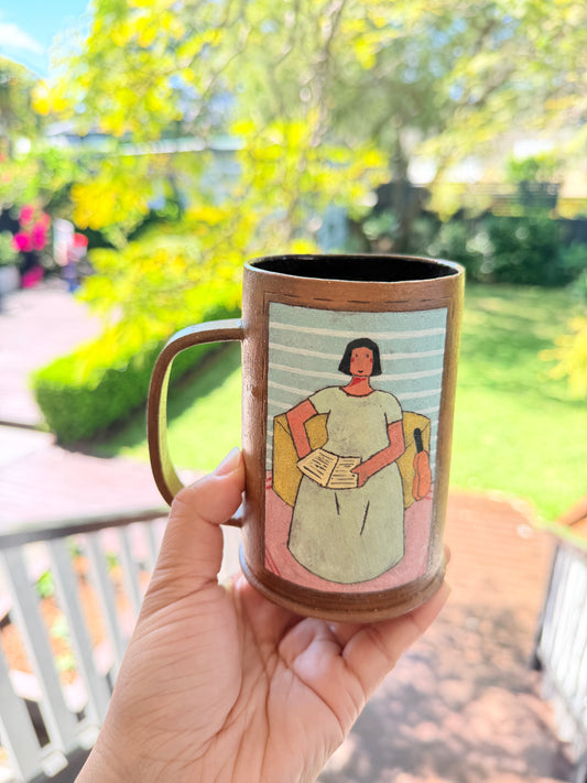 A hand holds a brown ceramic mug with a handle, featuring a painted illustration of a woman in a green dress holding a book, against a blurred outdoor garden background.