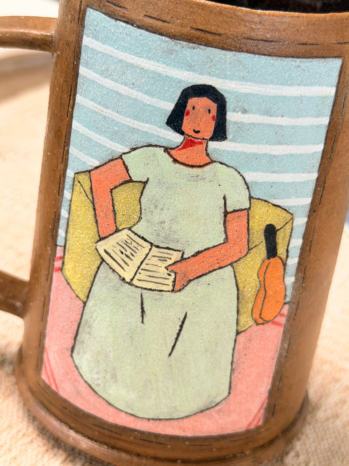 A close-up of a ceramic mug featuring a hand-painted illustration of a woman in a light green dress sitting in a yellow armchair and reading, set against a blue and white striped background.