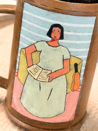 A close-up of a ceramic mug featuring a hand-painted illustration of a woman in a light green dress sitting in a yellow armchair and reading, set against a blue and white striped background.