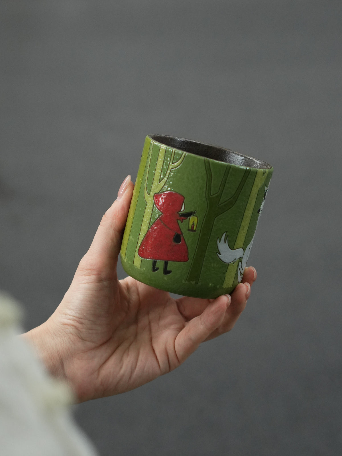 Hand holding a Little Red Riding Hood ceramic cup featuring the red-cloaked character carrying a lantern.