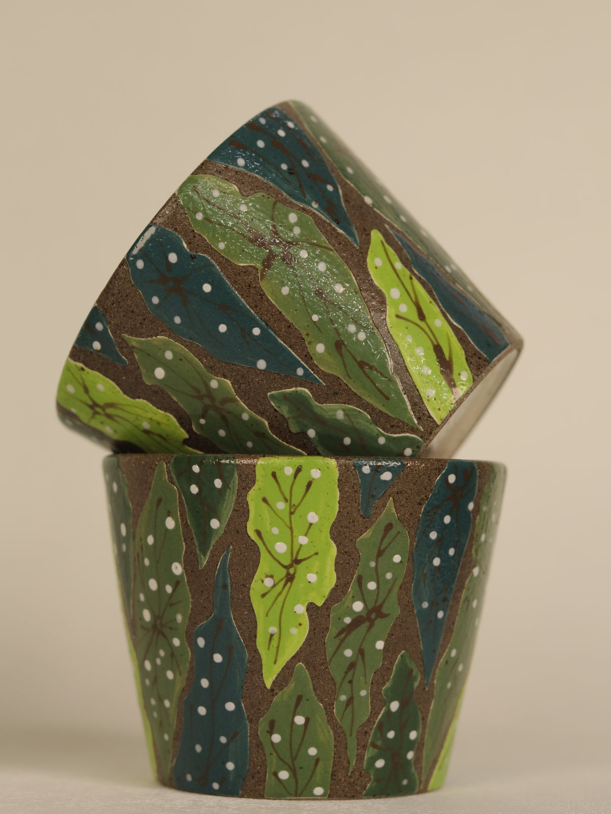Two stacked ceramic cups with a hand-painted forest pattern featuring green leaves, dark blue trees, and white dots on a brown background, set against a plain beige backdrop.