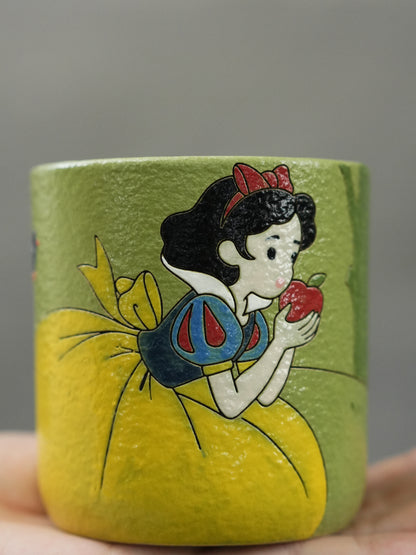 Hand holding a green ceramic cup featuring hand-painted Snow White with the enchanted apple.