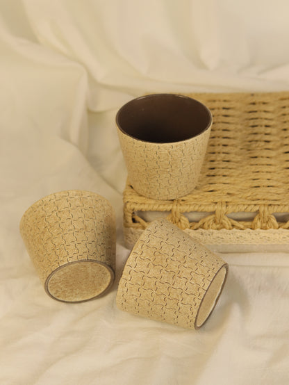 Three hand-carved geometric ceramic cups arranged on a woven tray and soft fabric.