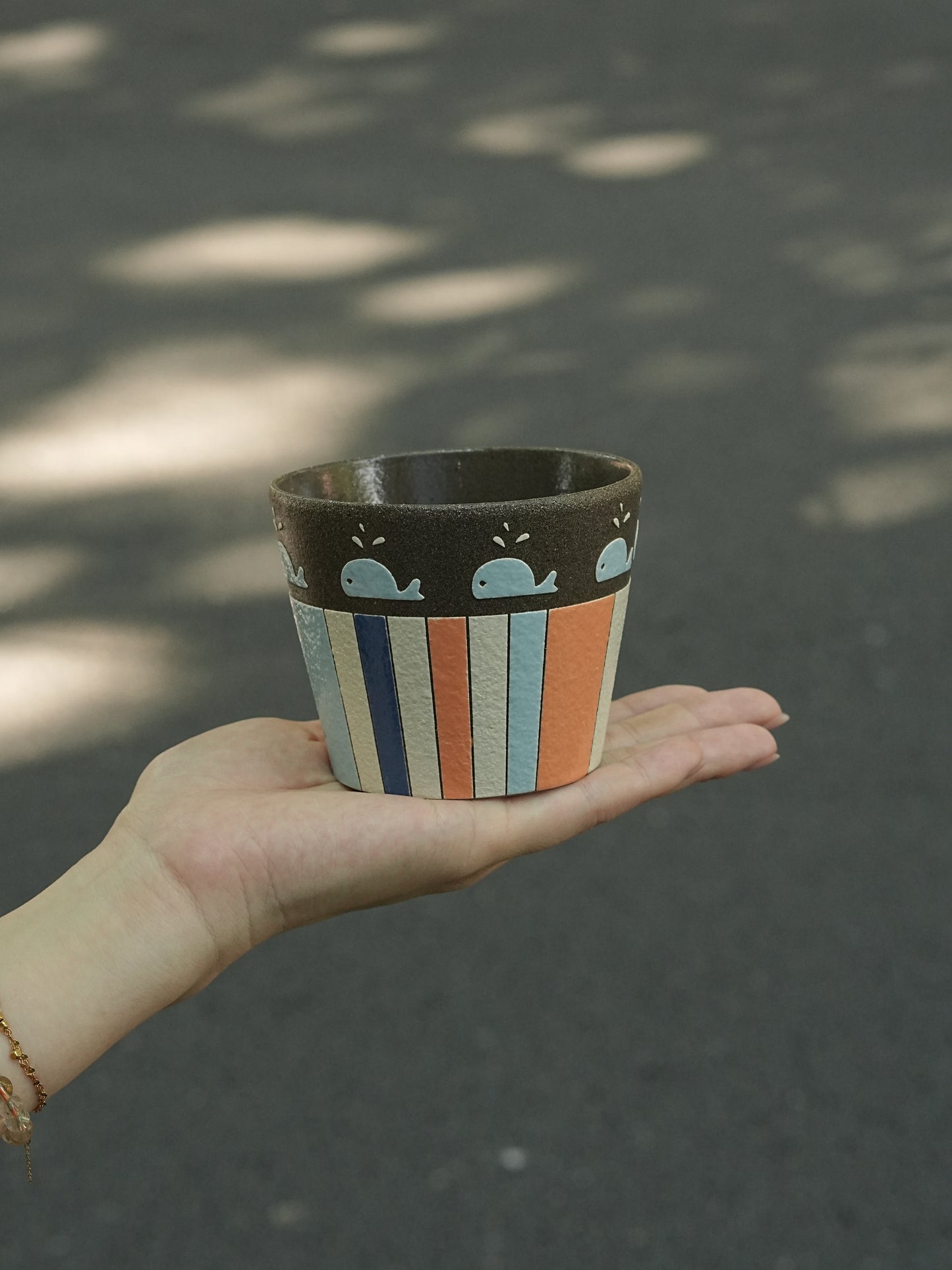 A hand holding a small ceramic cup outdoors on a paved surface with dappled sunlight and shadows.