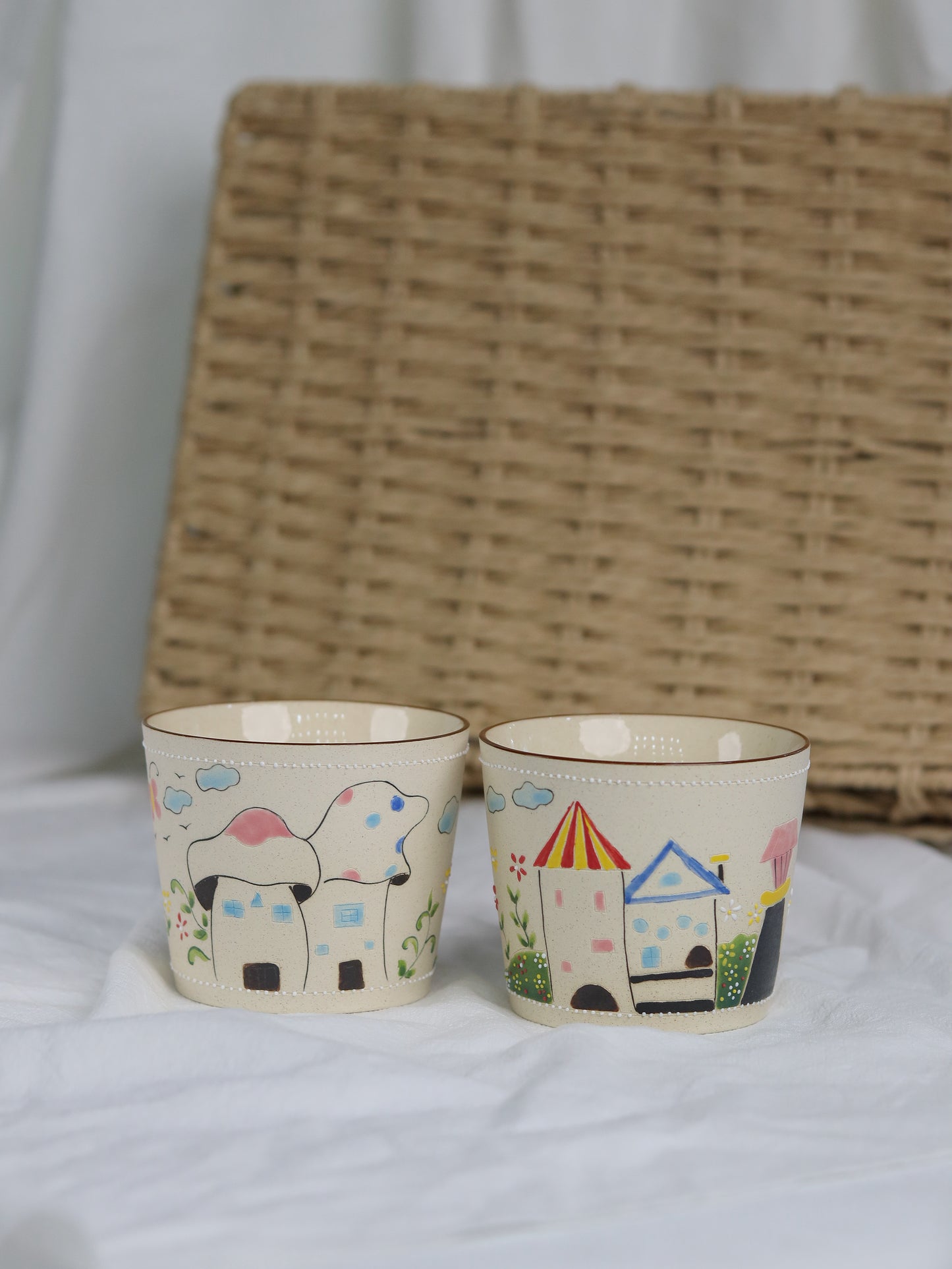 Two small cups with colorful illustrations of a house and a rainbow on a white surface with a woven basket in the background.