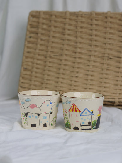 Two small cups with colorful illustrations of a house and a rainbow on a white surface with a woven basket in the background.