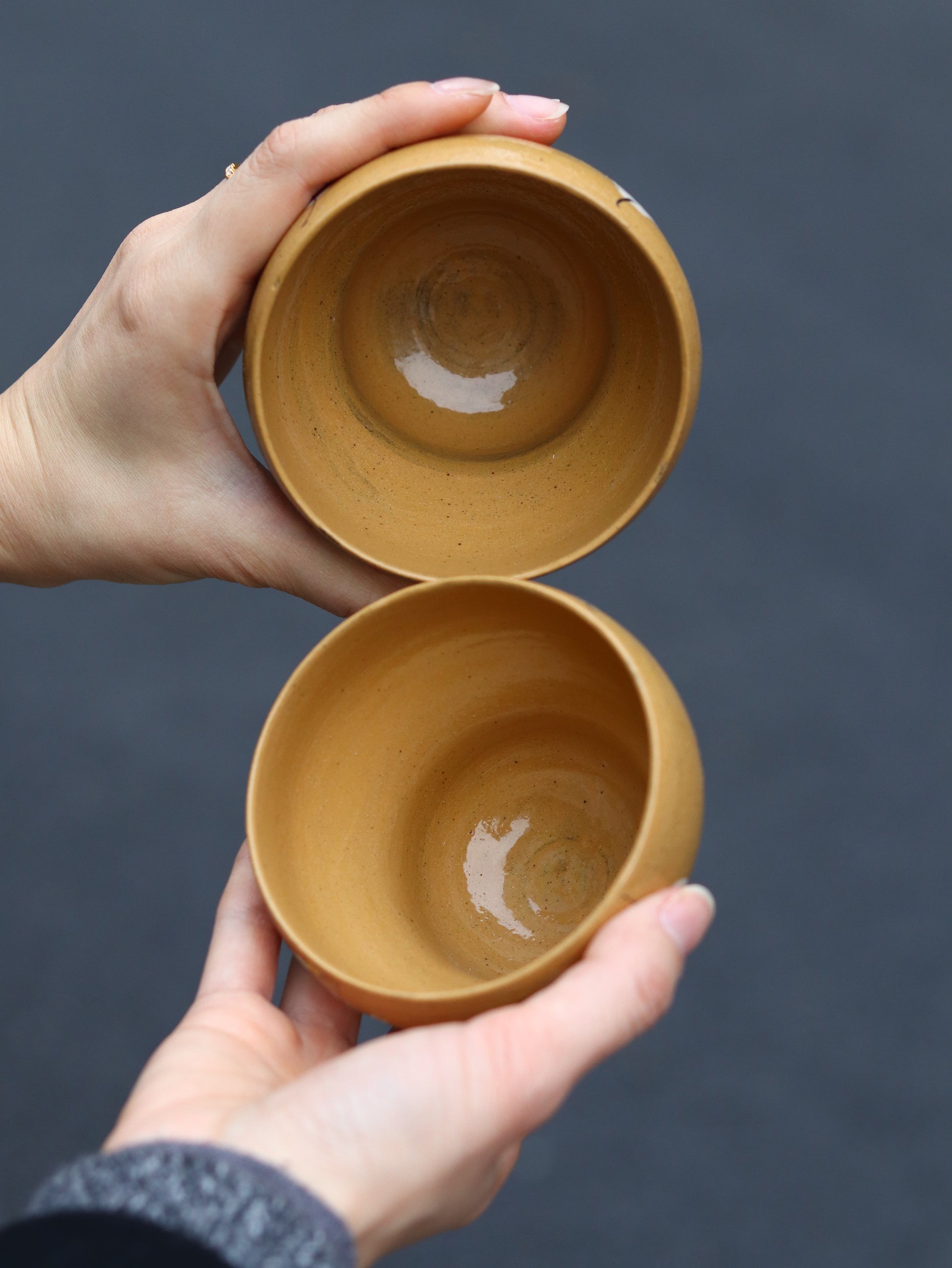 Two hands holding two small brown ceramic cups against a dark background